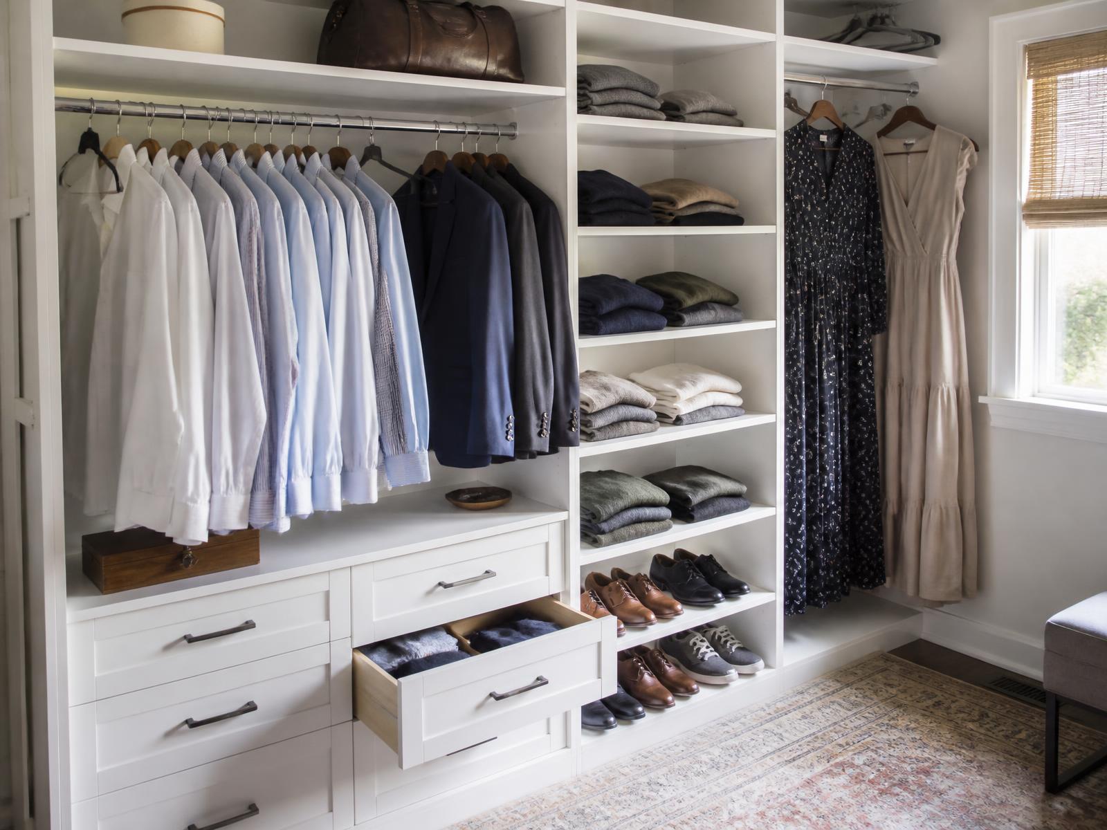 Walk-in closet with white cabinetry and black hardware holding white and blue dress shirts, navy and charcoal suits, folded sweaters, shoes and long dresses