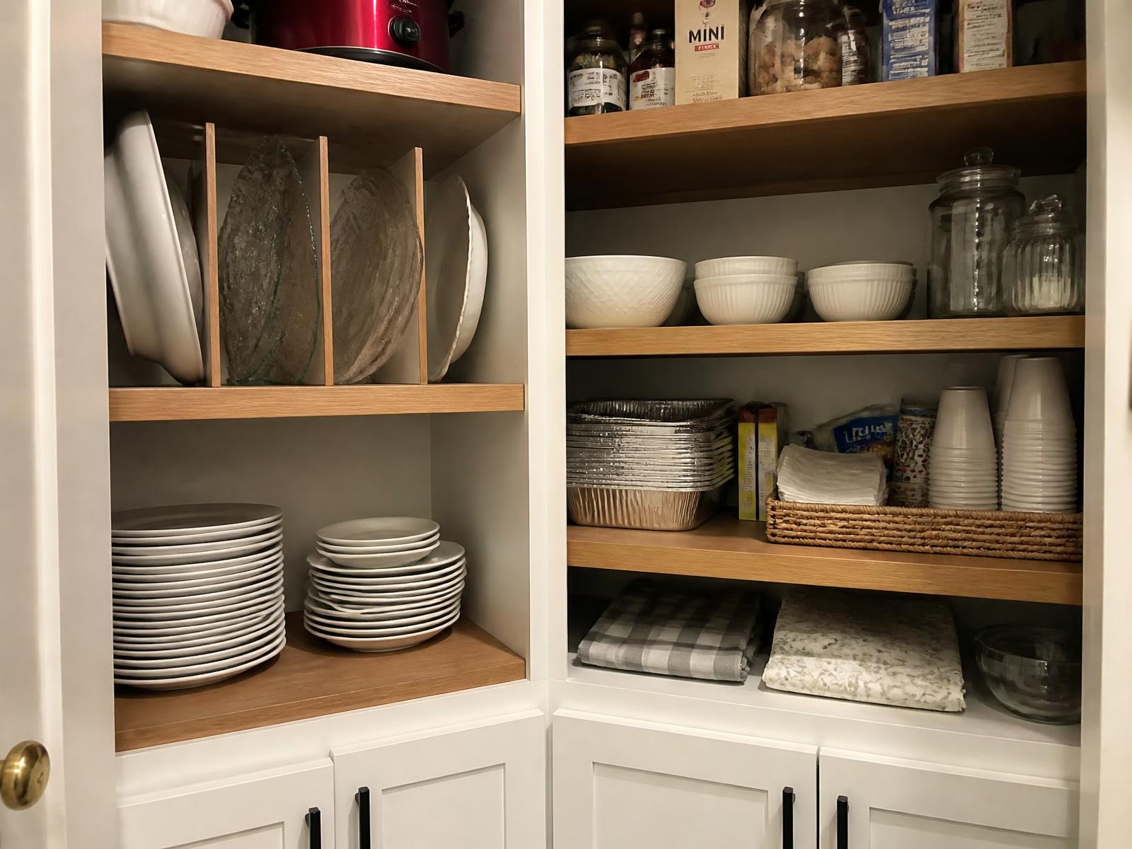 Pantry shelving with upright serving platters, stacked plates, bowls, foil pans, paper goods and folded tablecloths